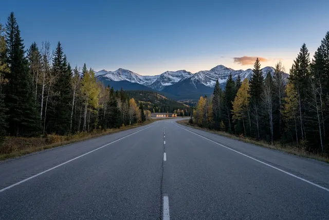 An empty two-lane highway running between spruce and yellow aspen toward snow-topped Rocky Mountain peaks at dusk, with the warm windows of a low building far ahead